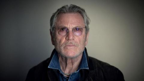 A photo of Tchéky Karyo, taken at La Baule film festival in July 2022 - he has grey hair, a beard and wears glasses and a denim shirt.