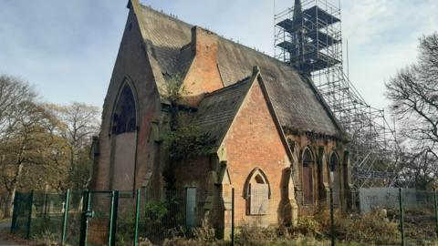 The dilapidated chapel is surrounded by a green fence and has boarded-up windows, as well as scaffolding to the back of the building 