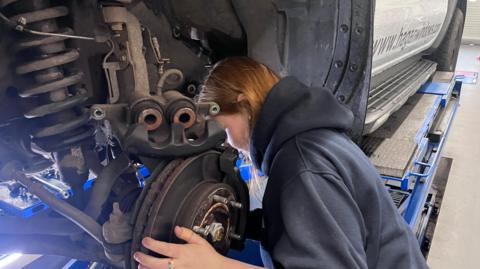 A close up of a female with ginger hair and a grey jumper who is replacing a car tyre.