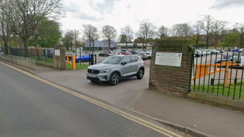 Street view shot of the car park. Several cars are already parked, while a grey SUV drives out of it, onto the main road.