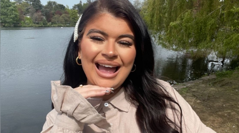 A woman is looking at the camera. She has one hand balanced on her chin and the other across her chest. She has a light cream top on and long dark hair. She is stood on the edge of a lake. She has an open mouth smile expression. 