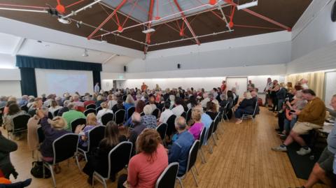 A large room with pale wooden floorboards and a high ceiling is filled with people sat on chairs attending the meeting, they are all facing away from the camera looking at a projector board 