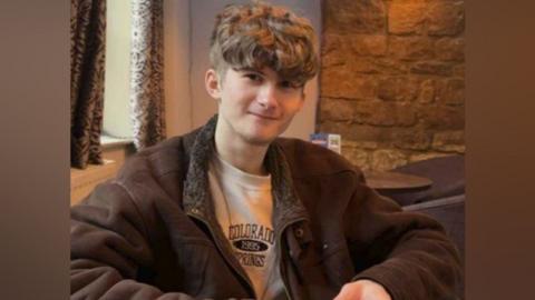 Cody Stock, wearing a brown jacket, cream jumper, sitting in a chair. He has short, curly, dark hair.
