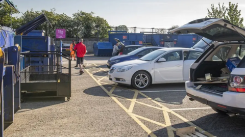An outdoor area with several large metal blue containers. There are parked cars, some with their boots open displaying rubbish. 