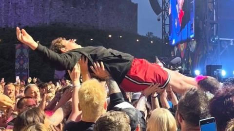 Jaret Reddick son, Everett Reddick doing a crowd surf during the Bowling for Soup show at Cardiff Castle. Everett is pictured being heled up by fans, with his arms in the air. He is wearing a black hoodie and red shorts. Three team members (two men and one woman) can be seen standing by as he is lifted.
