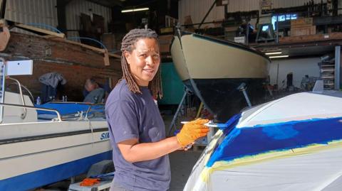 A woman stands in a boat yard building painting the hull of a small boat. She is smiling at the camera. She has short braids and is wearing a blue T-shirt. She is also wearing orange gloves. There are boats in the building behind her.
