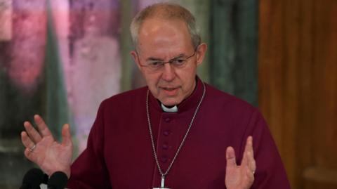 The Archbishop of Canterbury, dressed in a maroon cassock and a wearing a large silver cross on a chain, gestures with his hands as he speaks during the annual Lord Mayor's Banquet at Guildhall in London