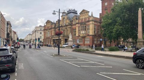 A high street with old buildings on either side, some cars parked on the left, showing and people crossing the road further down. 