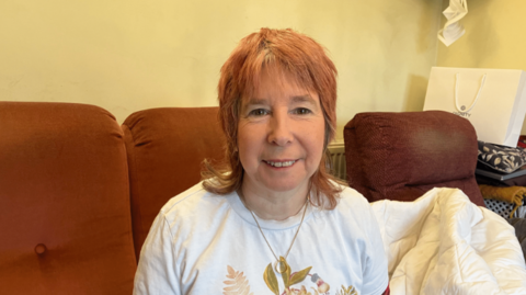 A woman with shoulder-length red hair wears a white top with a golden floral design on the front. She is pictured sitting on a dark orange sofa.
