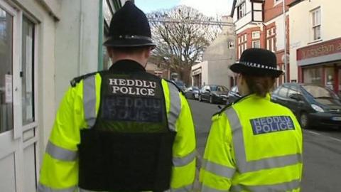 Police offers on the beat on a city street as seen from behind them. A male officer in a bobby hat with a high viz jacket and black gilet with the word police and the welsh translation in white letter. A woman officer, with a female style police hat is beside him  also in a high viz police jacket. 