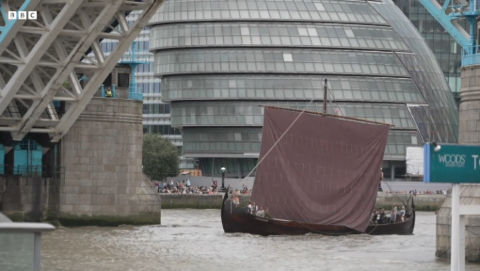 A Viking longship sails underneath London's Tower Bridge, on the River Thames. The boat is brown with a large, square red sail. The bridge is open, and people can be seen on the river bank in the background, in front of a large grey building.
