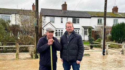 MP John Slinger wearing a dark jacket stood in flood water next to a man holding a broom and wearing a flat cap in front of a row of white houses