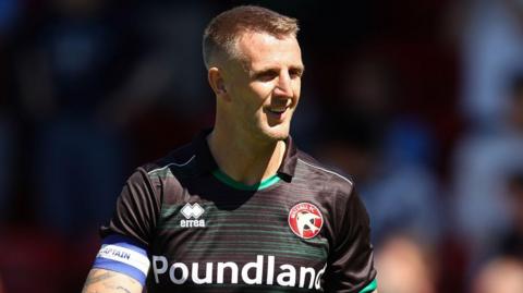 Peter Clarke wearing a black Walsall football top while on the pitch. He is squinting and smiling. He is looking away from the camera.