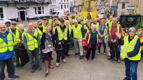 A group of people in hi-vis vests stand on a high street in Frome, all smiling at the camera, one woman at the front waving.