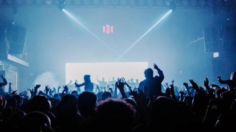A large silhouetted crowd of people dancing in front of the main stage at the old motion venue, which was inside a large warehouse. There are several people sitting on their friend's shoulders and everyone has their arms waving in the air. The room is smoky and and dark with a blue hue. 