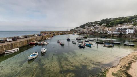 Small boats are moored in a harbour at low tide. One one side of the harbour there is a sea wall, with some cars parked on it. An opening is just visible towards the far right. There are houses and other low level buildings in  the top right hand side of the image and they are nestled besides the sea. There is a slight hill above them. Seaweed can be seen through the clear water in the harbour.