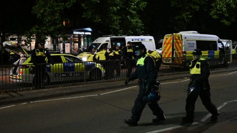 Two police officers wearing high-vis jackets walking across a road, with police cars, vans and further officers in the background