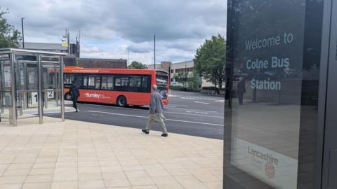 A view of the new bus station with a large flagged area in the foreground and large asphalt area with a red single decker bus driving past glass and chrome bus shelters. To the right is a digital sign saying 'welcome to Colne bus station'