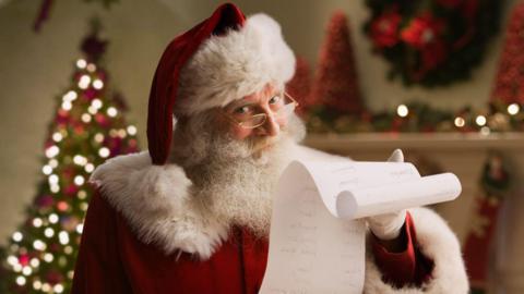 A man with a white beard and glasses wearing a red cloak and red hat with white fur trim, holding a long roll of paper with Christmas lights behind him