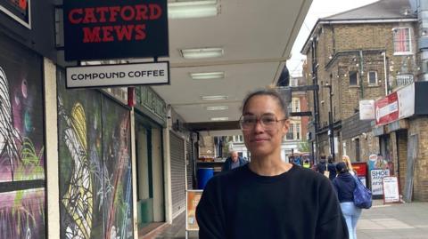 Gabriella is stood outside Catford Mews looking into the camera. She is wearing glasses and a black jumper. There is graffiti over the shutters of the cinema. 