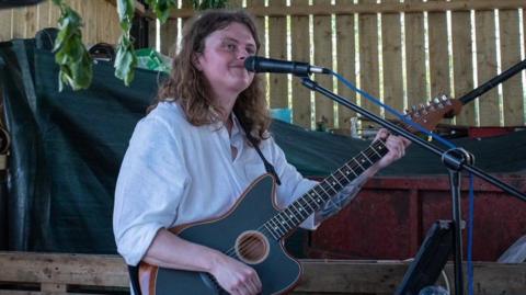 Ben Smith, who has shoulder-length hair and is wearing a white shirt, playing a guitar and singing into a microphone. He is smiling and looking into the distance. 