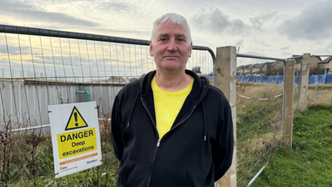 Keith Nairn, a man with white hair wearing a yellow t-shirt and black hoody stands in front of a metal fence