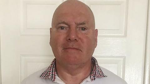 Philip Miller, who has a bald head and is wearing a white shirt with a dark red and black checked collar, is standing in front of a white door in a house. He looks impassive. 