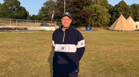 A man wearing a hat and quarter zip jumper standing in a green field with teepees behind him to his right and railings surrounding a burnt area on the grass to the left. Blue skies behind.