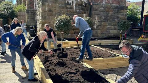 Nine men and women using gardening tools to fill raised wooden flower beds with soil. They are also adding plants. There is an old stone building in the background.