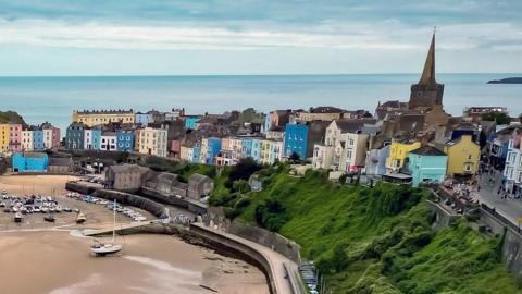 Aerial photo of the seaside town of Tenby in Pembrokeshire, Wales, showing rows of colourful houses overlooking the harbour with boats on the sand as the tide is out, with the Atlantic Ocean in the background behind the town's church spire and some clouds in the sky.