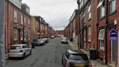 A residential street with back-to-back redbrick properties on both sides of the street. Parked cars and bins can also be seen.