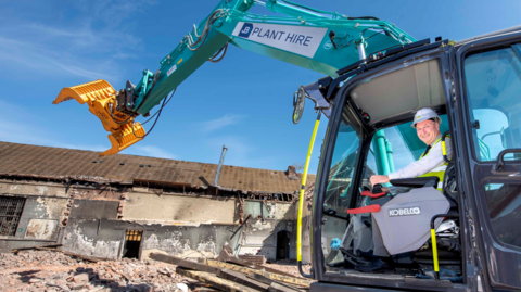 Steve Evans is sat in a digger that is being used in the building project. He is smiling at the camera as the building and rubble is visible on the left of the photo, as the building is demolished. 