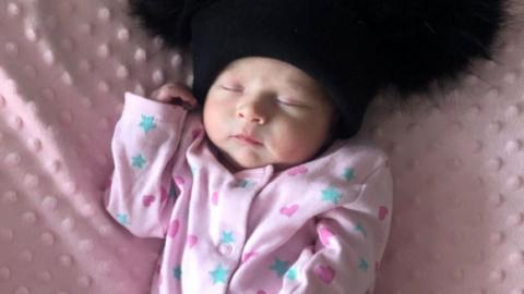A baby wearing a pink babygrow with blue stars and dark pint hears on it. She is asleep while lying on a pink blanket and wearing a black hat