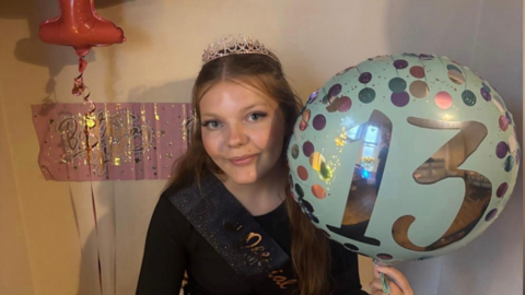 A young girl in a black dress and tiara and holding a balloon. She is celebrating her 13th birthday.