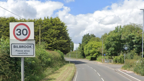 A Google street view image of the Bilbrook 30mph road sign, saying Please drive carefully, on Pendeford Mill Lane. Trees can be seen to the right and left of the road. 