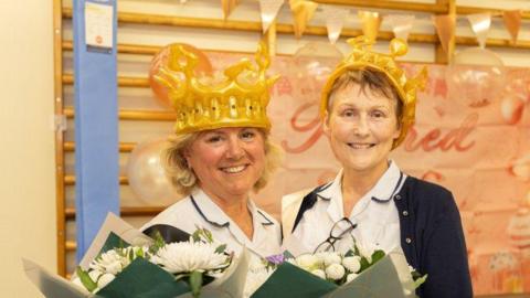 Two women stand in medical scrubs and smile at the camera while holding bouquets of white flowers. They both wear inflatable gold crowns.