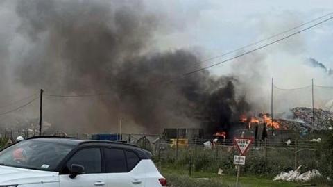 A view of a large smoke plume in the sky, flames at the bottom on large piles of rubbish. There are fences, a road and a couple of cars in the foreground.