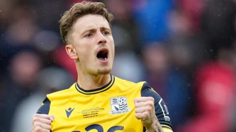 Harry Boyes has his mouth open towards the Southend fans after scoring a penalty at Wembley in the FA Trophy final with Wealdstone. He is wearing a yellow Southend away kit. 