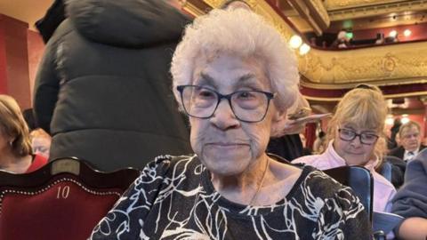 A woman with short white hair and glasses is seated in a red velvet theatre chair. She is wearing a black top with a white floral pattern, and the ornate theatre around her is filling with people.