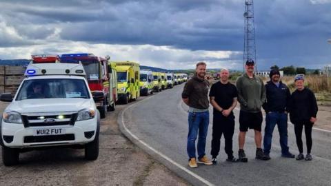 Five people stood on a road beside a row of parked emergency vehicles, with a large satellite dish on the horizon