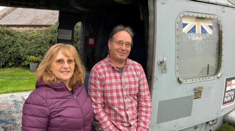 Man in a red check shirt and woman in a purple coat standing in front of a grey helicopter with curtains at the doors and a carpet