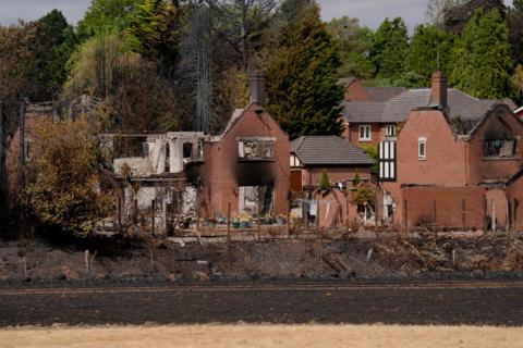 A row of burned out houses stand in front of trees.