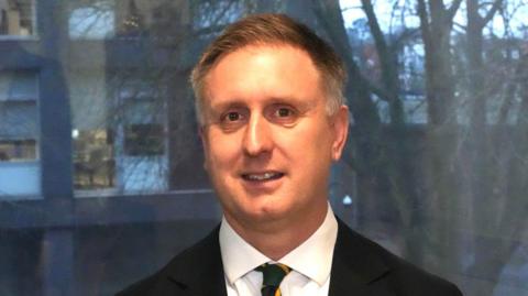 Ed Garrett smiles at the camera in front of a large window. He wears a black suit with a green and yellow tie. He has short grey hair.