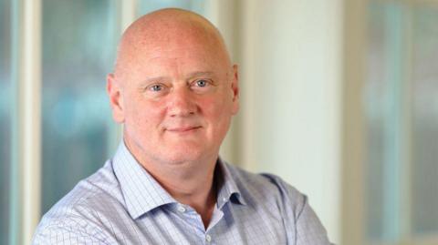 A bald man smiles at the camera. He wears a blue checked shirt. Blurred windows can be seen behind him.