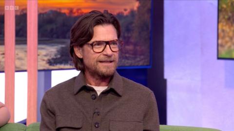 Jason Bateman has long hair swept back and wears glasses and a brown shirt