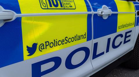 A close up of the exterior of a Police Scotland car. There are blocks of bright blue, yellow and white colours. The word "police" is in blue letters on a white background along the side of the car, and information on how to contact Police Scotland. 
