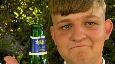 George Herd who has light brown hair cut very short at the back and in a long fringe on his forehead. He is wearing a grey jacket and a white shirt. He is holding up his right hand with an uncapped bottle and is also giving the thumbs up. Behind him are green trees and shrubs.
