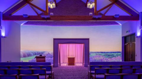 A purple landscape featuring numerous lavender bushes is displayed on a large canvas at the front of the chapel. In the middle of the chapel, there is a wooden coffin, and to the front there are four rows of benches. 