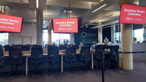 A meeting room with a large, long light wood table. There are black wheely chairs around them. There are several television screens displaying red backgrounds. Text on them reads "boundary review committee". There are people sitting further away from the camera, and a person standing up