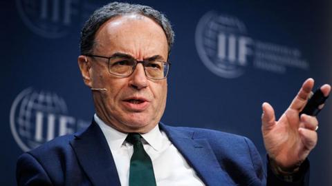 Andrew Bailey speaking on stage while sitting down at an International Institute of Finance event, the logo of which is visible on a backdrop behind Bailey. The governor has a serious expression, is mid-saying something, has a head mic, and is wearing a dark green tie with a blue jacket and white shirt. He is gesturing with his left hand as he speaks.